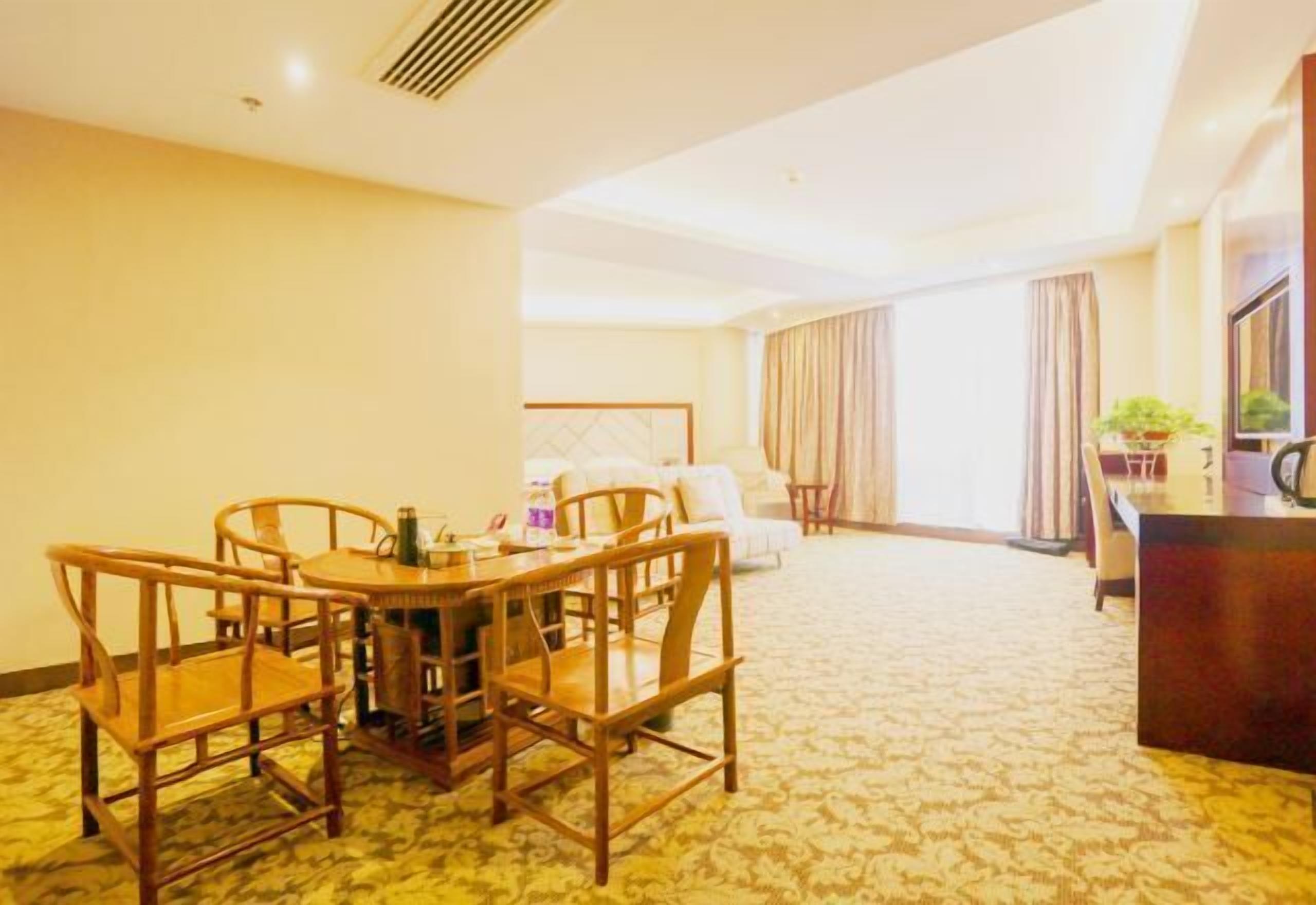 Habitación de hotel con comedor que incluye una mesa y sillas de madera, una cama con cabecera, un escritorio con silla y un ventanal con cortinas.
