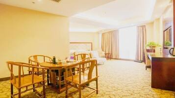 Habitación de hotel con comedor que incluye una mesa y sillas de madera, una cama con cabecera, un escritorio con silla y un ventanal con cortinas.
