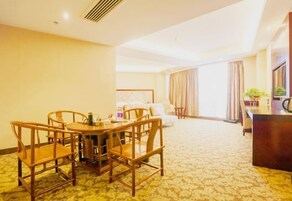 Habitación de hotel con comedor que incluye una mesa y sillas de madera, una cama con cabecera, un escritorio con silla y un ventanal con cortinas.