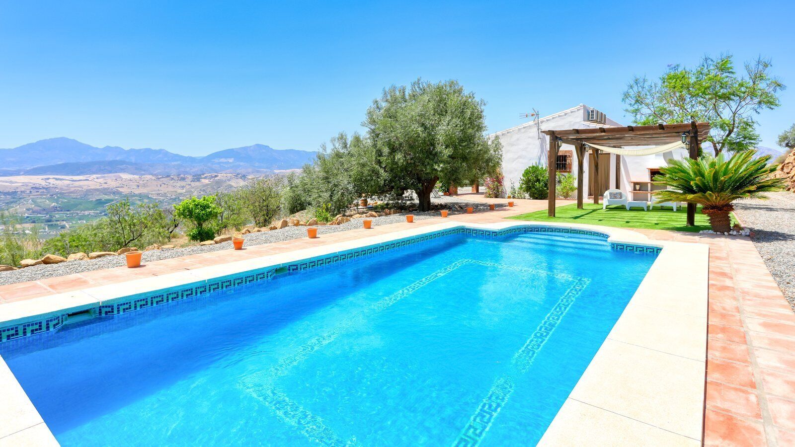 Una piscina rettangolare con bordi piastrellati, circondata da un patio con mobili da esterno e piante in vaso.
