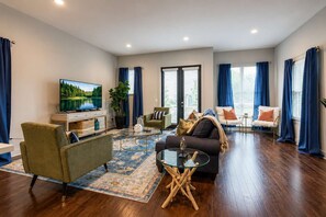 A modern living room with a flat-screen TV, comfortable seating, a coffee table, and a large window with curtains.