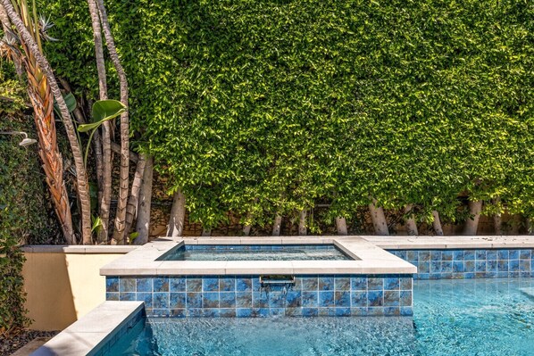 A swimming pool with a tiled edge, surrounded by a lush green hedge and tall palm trees.