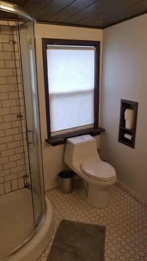 A bathroom with a glass shower enclosure, a toilet, a window with blinds, and a small shelf with toilet paper. - Fully Furnished Studio, Private Parking, Laundry, Private Outdoor Space (Venice)