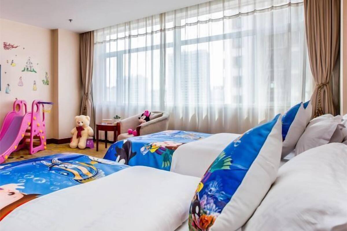 A hotel room with two beds, a desk, a chair, a teddy bear, a pink toy, and a window with curtains.
