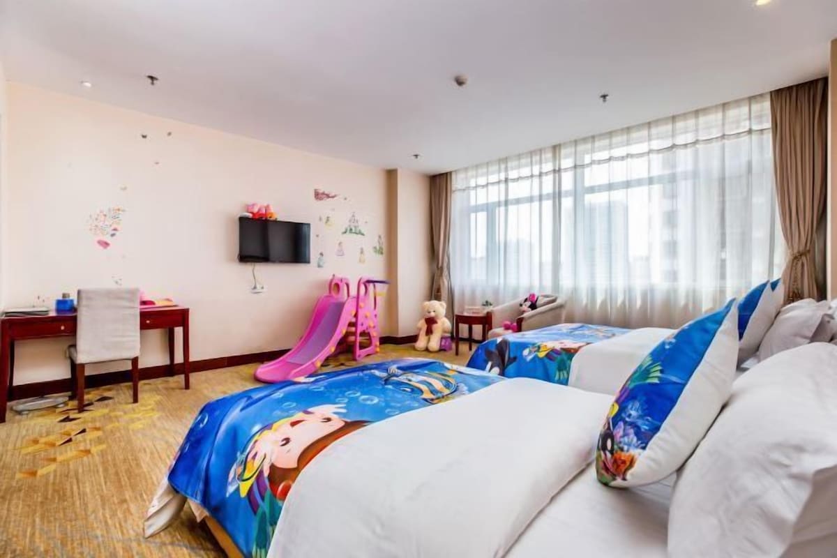 Habitación de hotel con dos camas, un escritorio, una silla, un televisor, una ventana con cortinas y una zona de juegos infantiles.