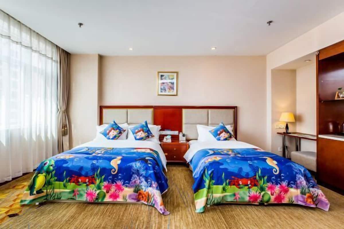A hotel room with two beds, each with colorful bedspreads, a wooden headboard, a nightstand, a lamp, and a framed picture on the wall.