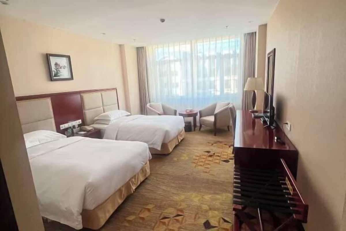 A hotel room with two beds, a desk with a lamp, a piano, and a large window with curtains.
