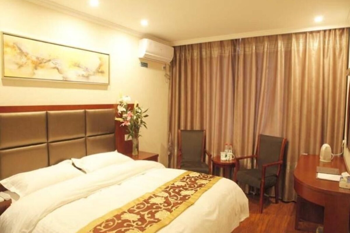 Quarto de hotel com cama, escrivaninha, duas cadeiras, mesa pequena com um vaso de flores e um quadro na parede.