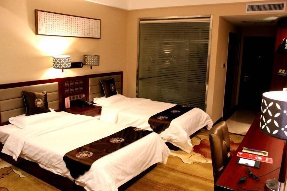 Habitación de hotel con dos camas, un escritorio y una silla. Cuenta con una ventana con persianas, una lámpara y un cuadro en la pared.
