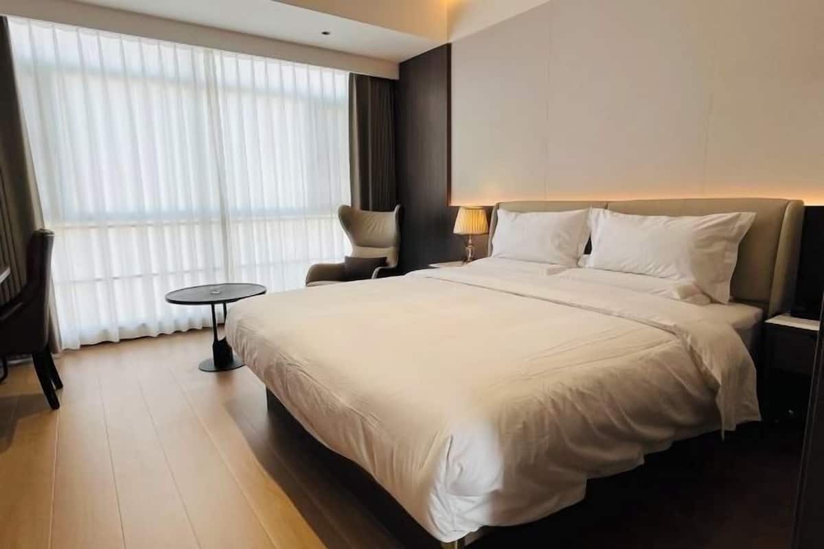 A hotel room with a large bed, a small round table, a chair, and a window with curtains.