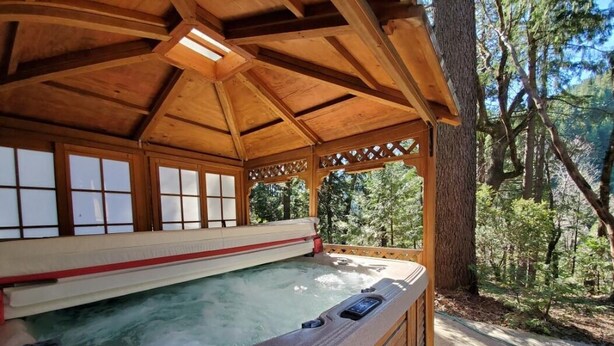 Ein hölzerner Whirlpool mit Glasverkleidung, umgeben von einer Holzterrasse und Bäumen.