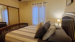 Ein Schlafzimmer mit einem Bett, einem Deckenventilator, einem Fenster mit Vorhängen und einem Nachttisch mit Lampe.