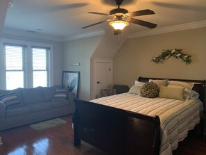 Une chambre à coucher comprenant un lit, un ventilateur de plafond, une fenêtre avec des stores, une porte et un élément de décoration fixé au mur.