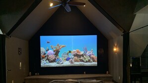 Un cinema en casa amb una pantalla gran que mostra una escena submarina, un ventilador de sostre i una zona d'seure acollidora amb coixins.
