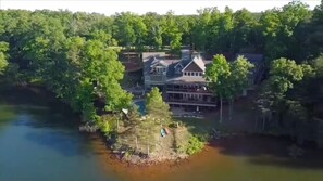 Vue aérienne d’une propriété au bord d’un lac, avec une grande maison entourée d’une forêt dense.