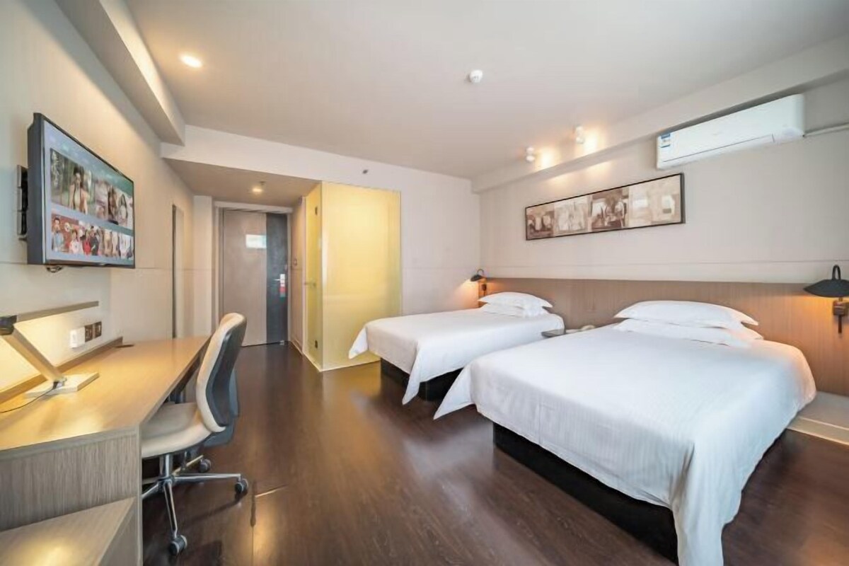 A hotel room with two beds, a desk, and a chair. There is a door leading to another room.