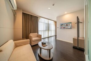 A modern hotel room with a sofa, a round coffee table, a large window with curtains, a painting on the wall, and a tall floor lamp.