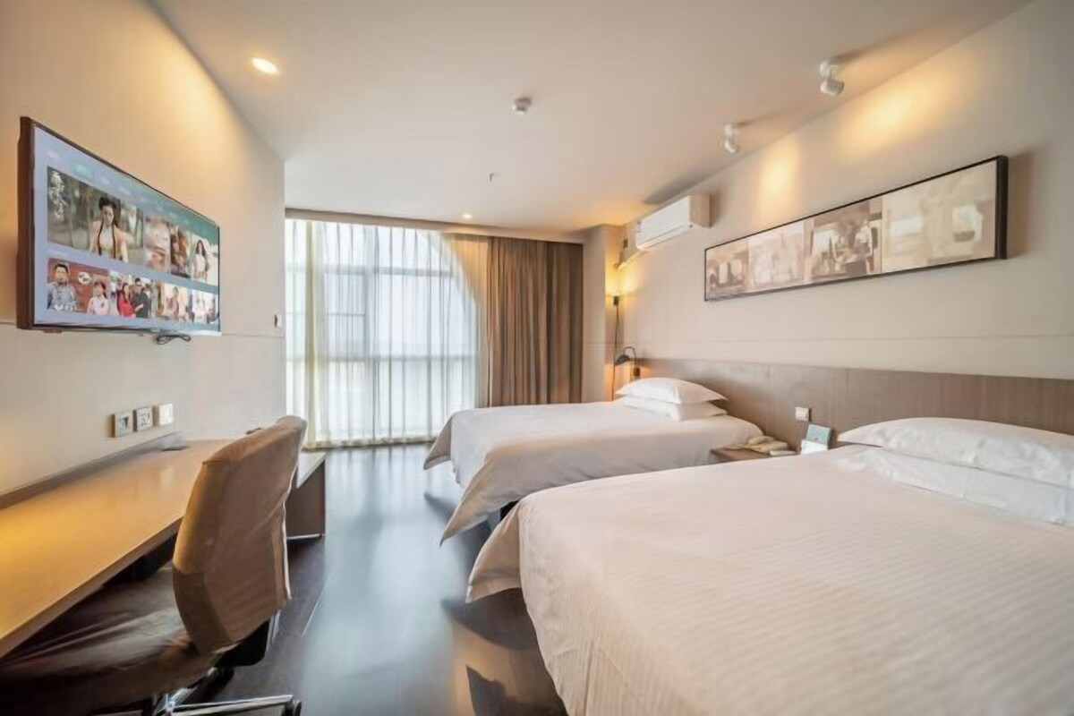 A hotel room with two beds, a desk, and a chair. There are large windows with curtains, and artwork on the walls.