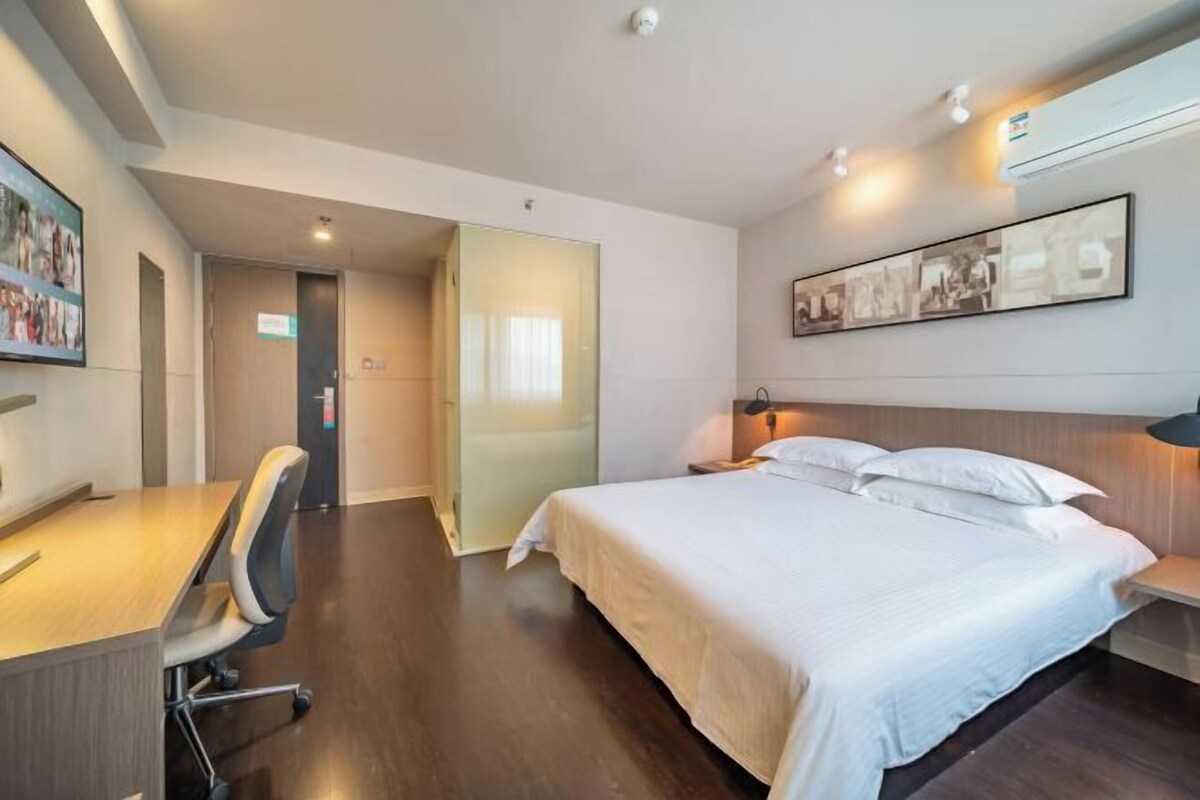 A hotel room with a bed, desk, chair, and a wall-mounted air conditioning unit.