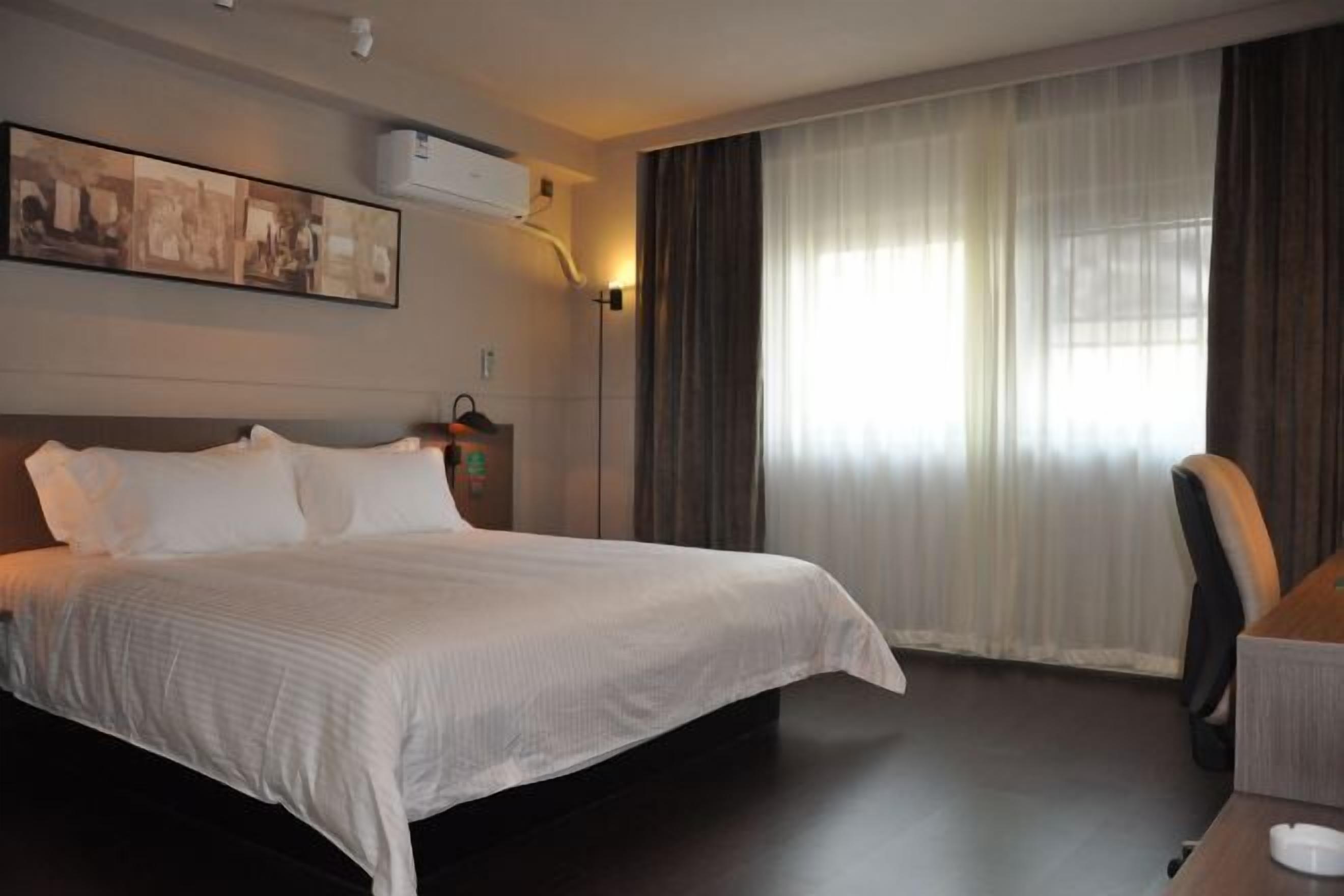 A hotel room with a bed, a desk, a chair, a painting on the wall, and a window with curtains.