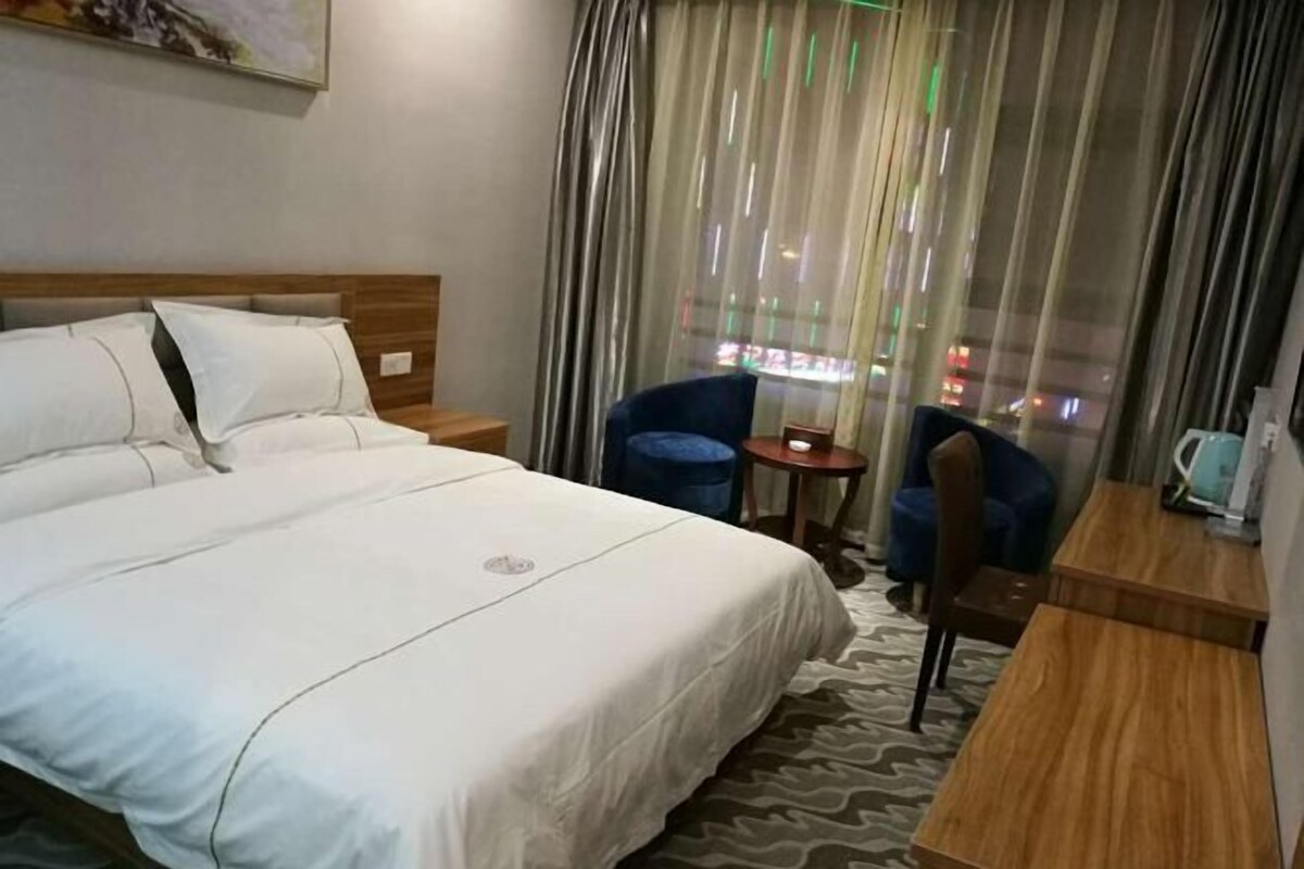 Ein Hotelzimmer mit einem großen Bett, einem kleinen Schreibtisch, einem Stuhl und Blick auf die Stadt durch transparente Vorhänge.