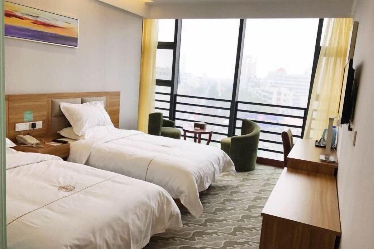 Ein Hotelzimmer mit zwei Betten, einem Schreibtisch, einem Stuhl, einem kleinen Tisch und einem großen Fenster mit Blick auf die Stadt.