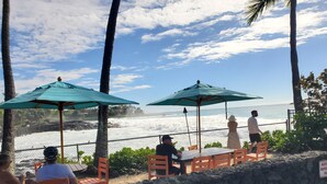 A beachside seating area with tables and chairs under umbrellas, overlooking the ocean.
