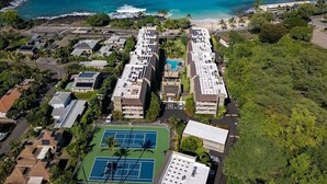 An aerial view of a resort with multiple buildings, a tennis court, and a beach in the background.