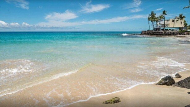 A beach with clear turquoise water, a sandy shore, and a building with palm trees in the background.