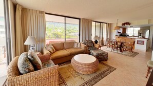 A spacious living area with a sofa, ottoman, and wicker chairs, a dining area with a table and chairs, and a kitchen visible through an open sliding door.