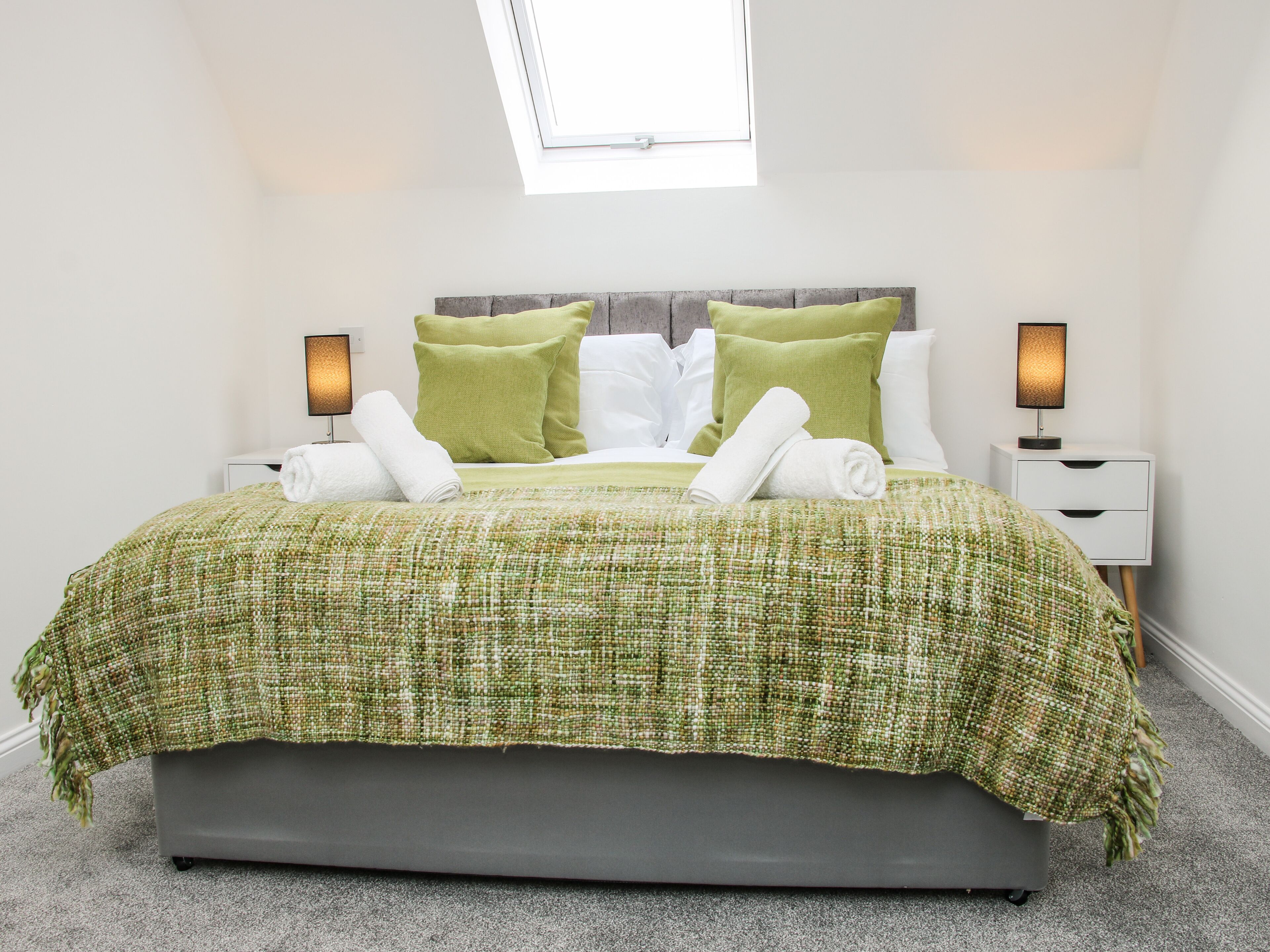 A neatly made bed with green and white bedding, a grey headboard, and two bedside lamps.