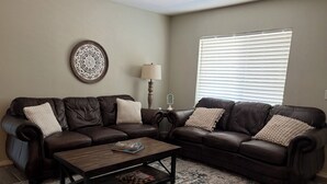 A living room with a brown leather sofa, a wooden coffee table, a patterned rug, a wall clock, and a window with blinds. - Newly remodel home 2BD/2BATH, 15min to dwtn Tulsa. (Owasso)
