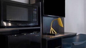 A modern kitchen with a black microwave, a refrigerator, and a flat-screen TV mounted above a desk with a yellow lamp. - Motel 6 Bonne Terre, MO (Bonne Terre)