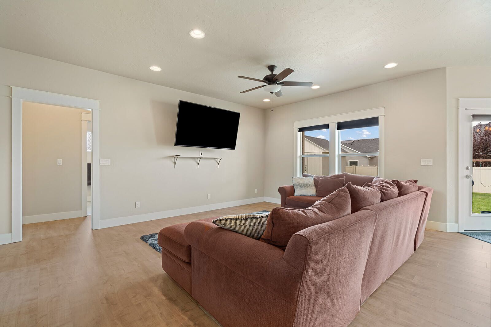 A modern living room with a large flat-screen TV mounted on the wall, a ceiling fan, and a sectional sofa.