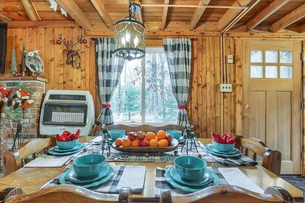 Comedor de una cabaña de madera con una mesa grande dispuesta para cuatro personas, rodeado de cortinas escocesas y vistas a árboles.