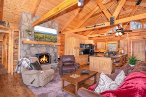 A cozy log cabin interior with a fireplace, comfortable seating, a kitchen area, and a stone wall.