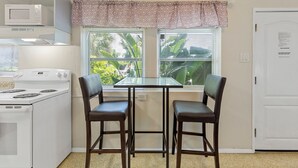 Une cuisine compacte équipée d’un four à micro-ondes, d’une cuisinière, de deux tabourets de bar, d’une petite table et d’une fenêtre avec des stores.
