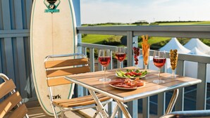 A table with a surfboard, wine glasses, and a plate of food on a balcony with a view of a green landscape.