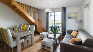 A modern living room with a wooden staircase, a dining table with chairs, a sofa, and a view of the outdoors. - Endroit Super! Appart avec Balcon | Local à Vélos (Biscarrosse)