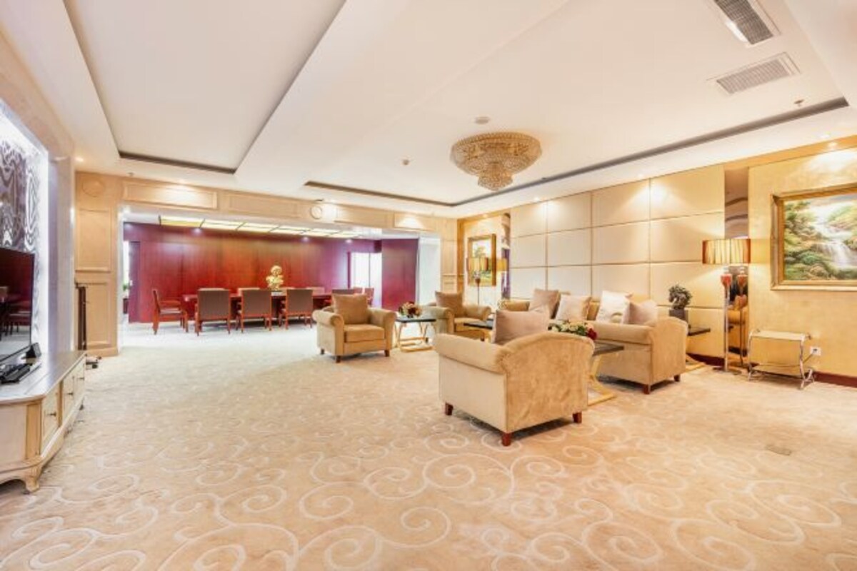 A spacious hotel lobby with a patterned carpet, plush seating, a reception desk, and a large painting on the wall.