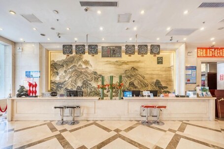 A hotel reception area with a traditional mural, a reception desk, two bar stools, and a clock.. Jinyuan Hotel