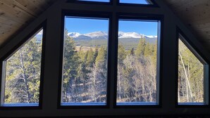 Huone, jossa on kalteva katto ja suuret ikkunat, joista avautuu näkymä lumihuippuisiin vuoriin ja metsäiseen maisemaan.