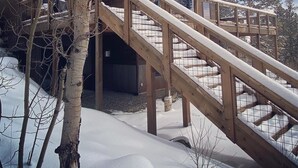 Puinen mökki, jossa on puinen portaikko, joka johtaa terassille. Mökkiä ympäröi lumi ja puut.