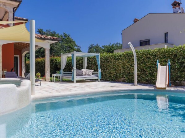 A clear blue pool with a white canopy and a slide, surrounded by a patio area with seating and a bed.