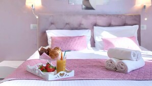 Een hotelkamer met een bed, roze kussens, een dienblad met croissants en aardbeien, een opgerolde handdoek, en een schilderij van roze bloemen aan de muur.
