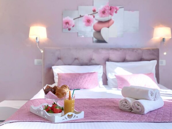 A hotel room with a bed, pink pillows, a tray with croissants and strawberries, a rolled towel, and a painting of pink flowers on the wall.