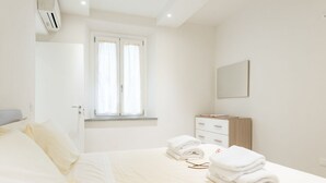Ein Hotelzimmer mit Bett, einem Fenster mit Vorhängen, einem Nachttisch mit Lampe und einem an der Wand befestigten Spiegel.