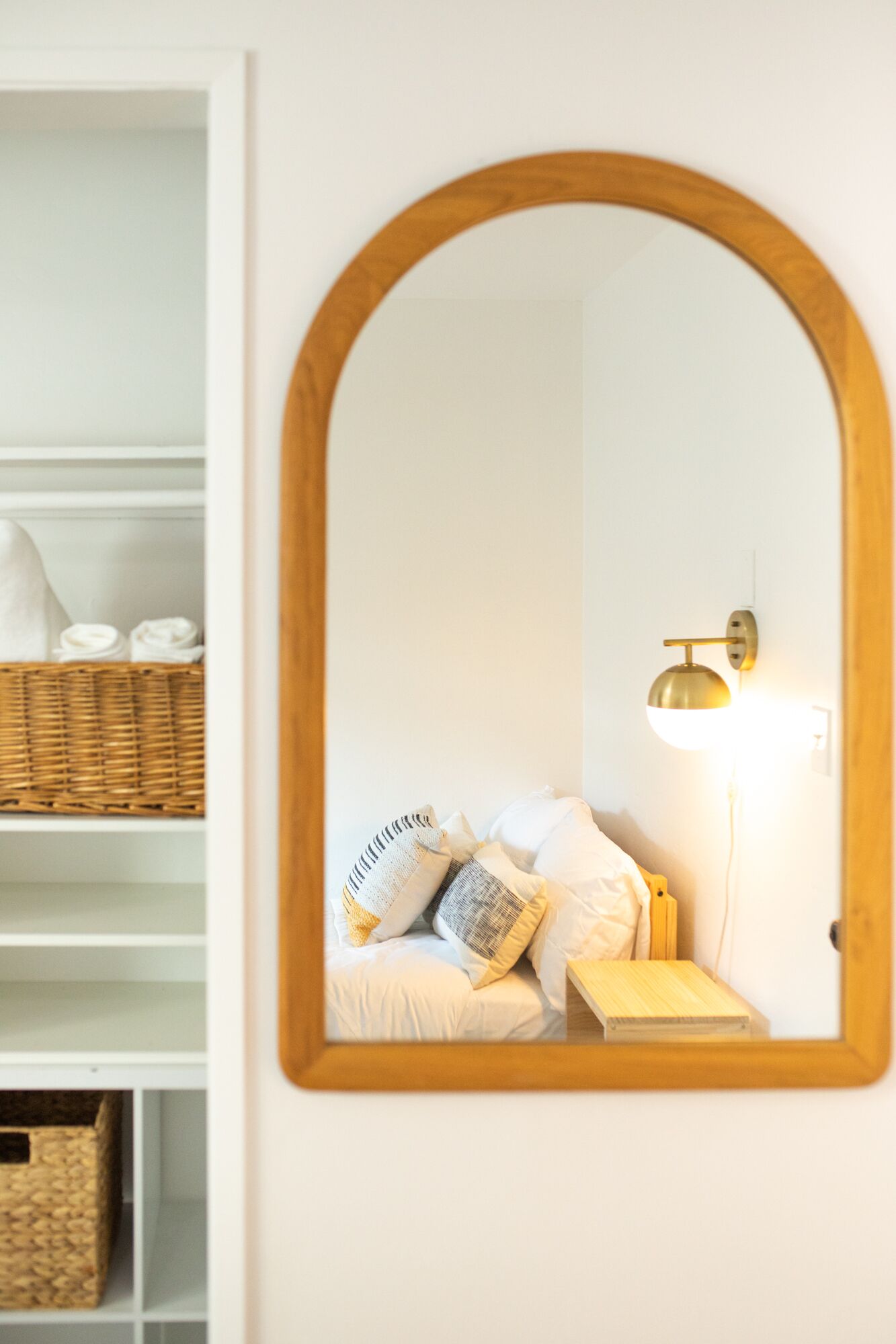 Una camera da letto con uno specchio con cornice in legno, una lampada da comodino, un letto con biancheria bianca e un cesto in vimini su uno scaffale bianco.
