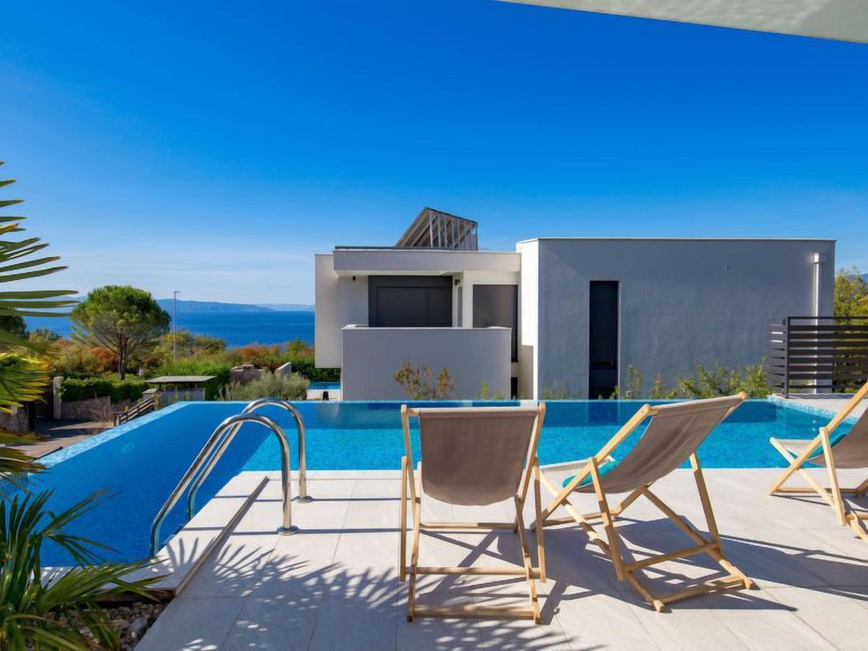 A modern pool area with a clear view of the sea, a contemporary building, and two wooden lounge chairs.