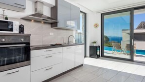 A modern kitchen with a built-in oven, microwave, and stove, a large window with a view of the sea, and a balcony with a pool and lounge chairs. - Apartment Albi by Interhome (Rijeka)